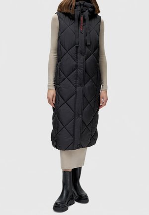 Femme portant un long gilet matelassé noir sans manches sur une robe beige et des bottes noires épaisses, debout devant un fond uni.