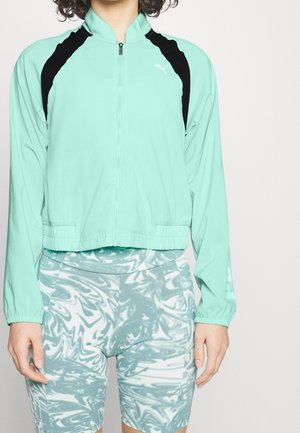 Kvinde iført en mintgrøn atletisk jakke med lynlås og sorte detaljer samt højtaljede cykelshorts i blå og hvid marmoreret mønster.