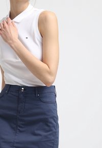 Vit ärmlös polo med krage och en liten logotyp, kombinerad med en mörkblå denimkjol med framfickor och dragkedja.