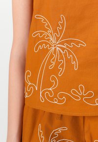 Braunes Baumwollträgertop mit weißem, gesticktem Palmenmotiv und geschwungenen Mustern am unteren Saum. Glatte Textur, lässige Passform.