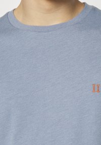 Light blue cotton crew neck t-shirt with short sleeves, featuring an orange embroidered "II" accent on the chest. Soft texture and smooth finish.