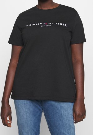 Persoon draagt een zwart Tommy Hilfiger T-shirt met logo en 'EST. 1985' gedrukt op de borst, gecombineerd met een blauwe spijkerbroek.