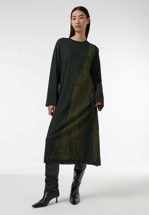 Grünes Langarmkleid mit einem vertikalen Muster aus gelben Linien. Hergestellt aus weichem Stoff, mit einem A-Linien-Schnitt und entspannter Passform. Kombiniert mit schwarzen kniehohen Stiefeln.