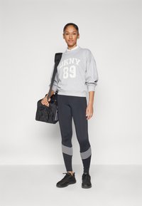 Pilkas megztinis su baltu spaudiniu "OKNY 89", juodos leginsai su pilkomis akcentais ir juodi sportbačiai. Modelis laiko juodą sportinį krepšį.