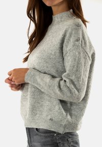 Maglione lavorato a maglia grigio chiaro con un motivo a rombi testurizzato, collo a coste e bottoni decorativi sulla spalla. Materiale morbido, vestibilità comoda.