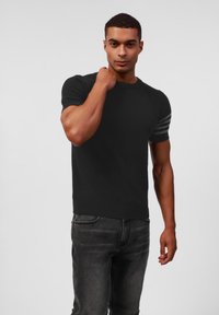 Svart kortermet t-skjorte med ribbestruktur og tre hvite horisontale striper på den ene skulderen, kombinert med mørk grå jeans.