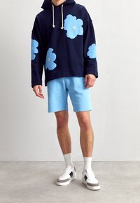 Felpa con cappuccio blu navy con ampi motivi floreali blu, dettagli beige, abbinata a pantaloncini azzurri. Scarpe da ginnastica bianche con dettagli grigi e blu navy.