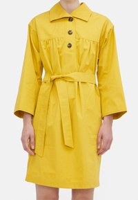 Abito di cotone giallo con colletto, tre bottoni e vita con cintura; presenta maniche lunghe e dettagli arricciati sul davanti.