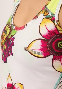 Kukallinen toppi, jossa on kirkkaan pinkkejä ja keltaisia kukkia valkoisella taustalla, ja värikäs etiketti näkyvästi kankaan päällä.