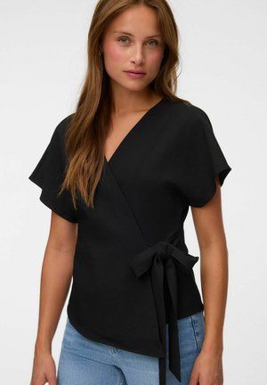 Femme aux longs cheveux bruns portant un haut cache-cœur noir à manches courtes noué à la taille et un jean bleu clair, sur un fond clair uni.