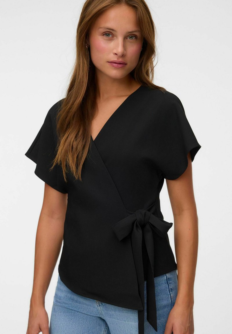 Femme aux longs cheveux bruns portant un haut cache-cœur noir à manches courtes noué à la taille et un jean bleu clair, sur un fond clair uni.