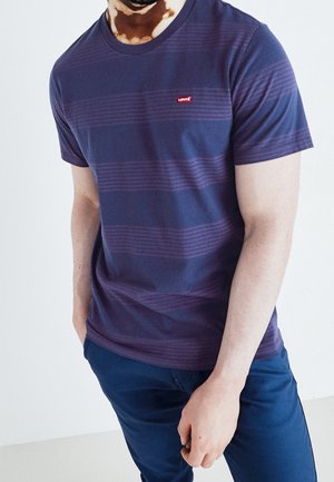 Mand iført en navyblå stribet Levi's t-shirt og navyblå bukser, med den ene hånd i lommen mod en ensfarvet hvid baggrund.