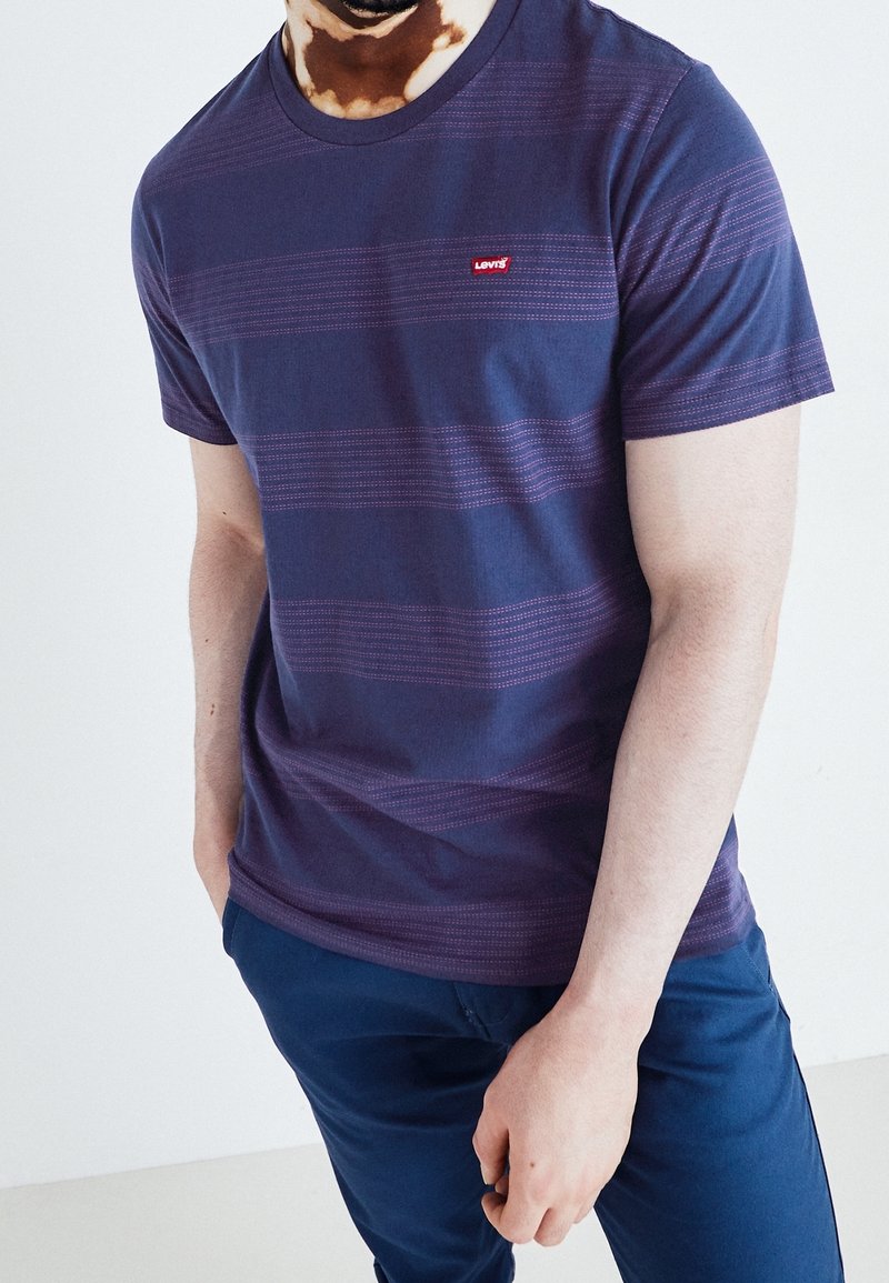 Homme portant un t-shirt rayé bleu marine Levi's et un pantalon bleu marine, une main dans la poche, sur un fond blanc uni.