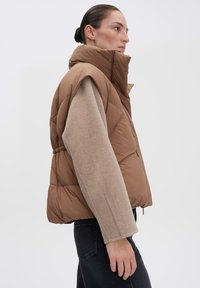 Braune Weste mit Stehkragen, gepolstert und mit Kordelzug in der Taille. Darunter trägt die Person einen hellbeigen Pullover mit langen Ärmeln.