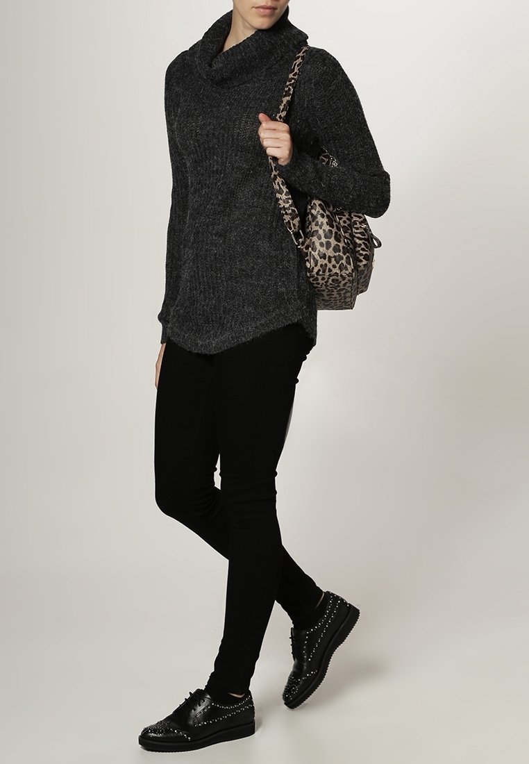 Personne portant un pull à col roulé gris foncé, un pantalon noir, des chaussures noires cloutées, et un sac à dos imprimé léopard porté sur une épaule.