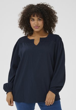 Navy blauwe blouse met een V-hals en smockdetails op de schouders. Lange, volumineuze mouwen met elastische manchetten en een relaxte pasvorm.