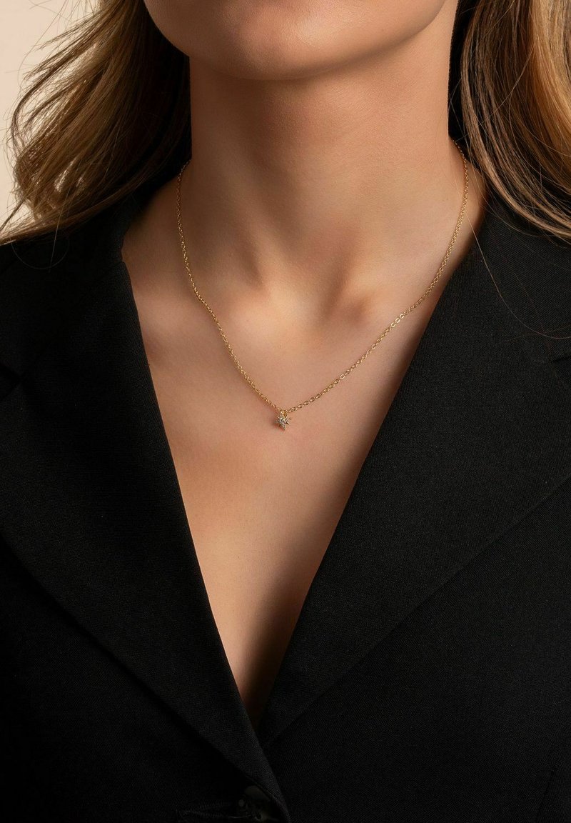 Guldkedja med en liten stjärnformad berlock, bärs på en naken hals, mot en svart tygbakgrund.