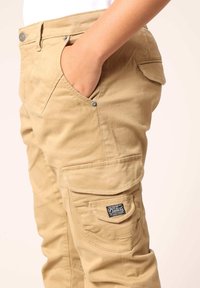 Beige cargobroek van duurzaam stof, met meerdere zakken, een logo patch en metalen drukknoopdetails op de taille en zakflappen.