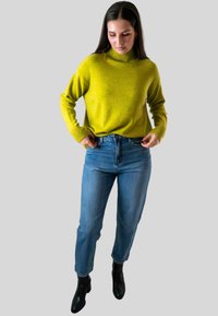 Vrouw die haar blauwe jeans aanpast, gekleed in een heldergeel coltrui en zwarte enkellaarsjes, staande tegen een eenvoudige lichte achtergrond.