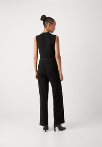 Combinaison noire sans manches avec un buste ajusté et des pantalons larges, accentuée d'une ligne de clous à la taille. Texture de tissu lisse.