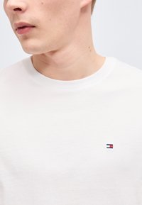 Νεαρός άνδρας που φοράει απλό λευκό μπλουζάκι με στρογγυλή λαιμόκοψη και μικρό κόκκινο, λευκό και μπλε λογότυπο στο αριστερό στήθος.