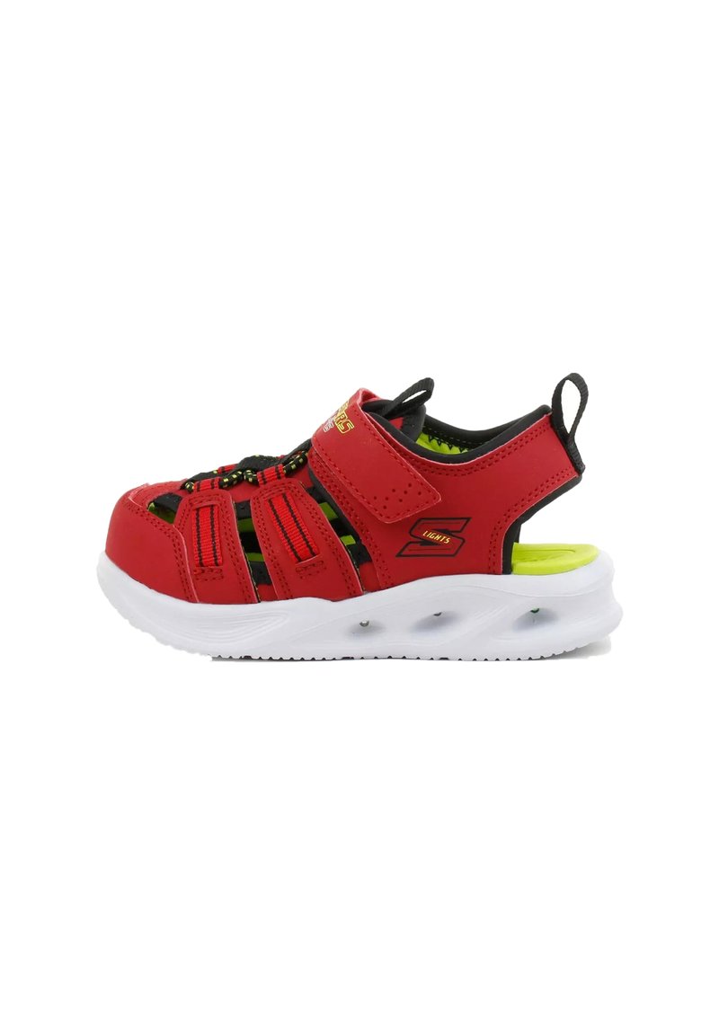 Zapatilla deportiva roja con una parte superior transpirable, correa ajustable, detalles en negro y una suela blanca acolchada. Presenta un forro interior y cordones verdes.