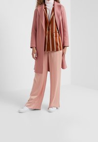 Růžový kabát přes hnědobílý pruhovaný blejzr, doplněný měkkými růžovými kalhotami s širokými nohavicemi a bílými teniskami. Všechny kusy mají hladkou texturu.