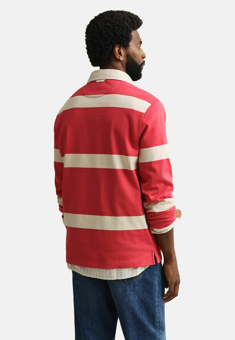 Mann mit lockigem Haar und Bart, der ein rot-beiges, gestreiftes Langarmshirt über einem karierten Hemd und blauen Jeans trägt, seitlich stehend mit entspannten Händen.