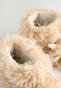 Miękkie beżowe, puszyste buciki dla niemowląt z fluffy teksturą, z małym zapięciem z materiału z jednej strony.