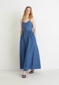 Robe maxi en denim avec des bretelles fines, fermeture à boutons sur le devant et taille froncée. La couleur est bleu moyen avec une texture lisse.
