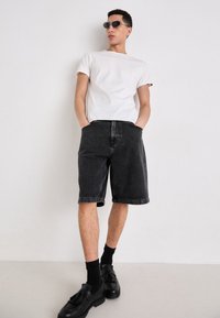 Schwarze Denim-Shorts, weißes T-Shirt und schwarze Slipper mit Quasten. Die Accessoires umfassen dunkle, runde Sonnenbrillen. Einfarbige Farben, lässige Passform.