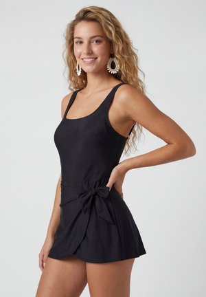 Femme aux longs cheveux blonds ondulés portant de grandes boucles d'oreilles blanches et une robe noire sans manches avec un nœud noué à la taille, souriante et posant.
