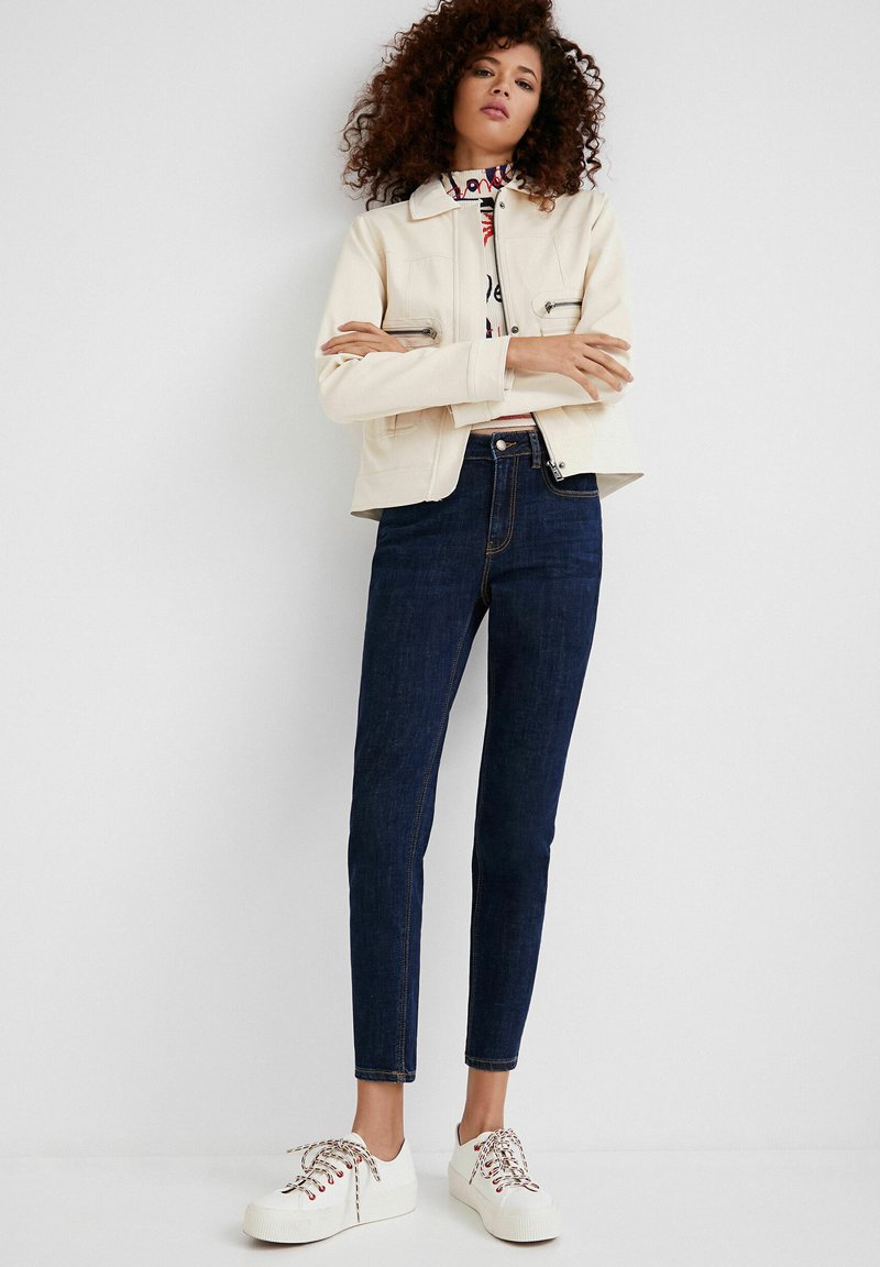 Beige cropped jack, witte gebloemde blouse, donkere skinny jeans en witte sneakers met meerkleurige veters. Eenvoudig ontwerp en strakke lijnen.