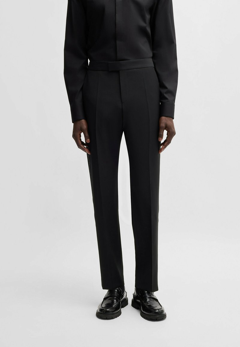 Eine Person trägt eine eng anliegende schwarze Anzughose, ein schwarzes, knopfbares Hemd und schwarze, polierte Slipper und steht vor einem schlichten hellen Hintergrund.