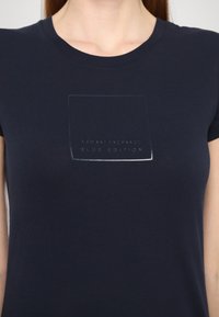 Maglietta blu navy realizzata in morbido cotone con collo a giro. Presenta una grafica del logo trasparente "AX" con "ARMANI EXCHANGE BLUE EDITION" sottostante.