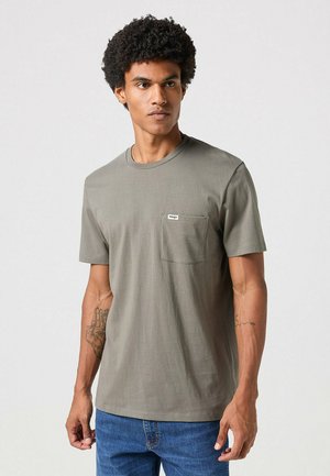 Camiseta de manga corta verde oliva hecha de algodón suave, con un bolsillo en el pecho que tiene una pequeña etiqueta. Jeans de mezclilla azul a juego visibles.