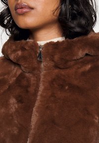 Femme portant une veste en fausse fourrure marron avec une fermeture éclair argentée partiellement fermée, montrant un haut crème en dessous.
