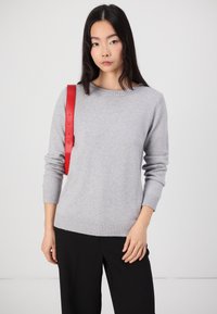 Hellgrauer Pullover mit langen Ärmeln und geripptem Saum, kombiniert mit schwarzen Hosen. Ein rotes Lederband liegt über einer Schulter.