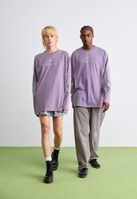 Chemises à manches longues violet avec "TEAMGEIST" imprimé, associées à un pantalon en velours côtelé gris et une jupe superposée à carreaux. Bottes de combat noires portées.