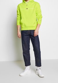 Felpa verde neon con lacci e tasca frontale, abbinata a pantaloni in denim scuro e sneakers bianche. Logo semplice visibile sulla felpa.