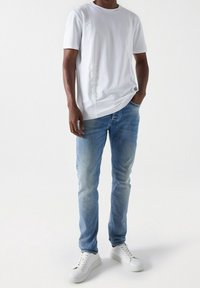 White short-sleeve t-shirt with subtle side seam details, paired with light blue denim jeans and white sneakers.
