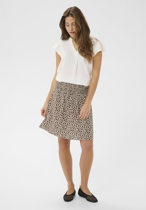 Vrouw staand, gekleed in een wit T-shirt met V-hals en korte mouwen, een rok met luipaardprint en zwarte gevlochten platte schoenen, tegen een eenvoudige lichte achtergrond.