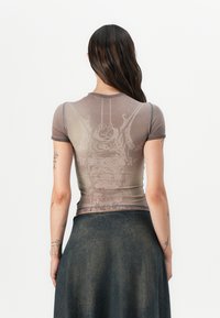 Kortsleeve, tettsittende brun mesh-top med et subtilt abstrakt mønster. Har en rund halslinning og kontrasterende sømmer langs kantene.