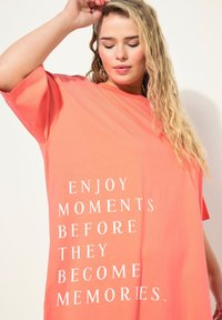 Camiseta oversize de coral hecha de algodón suave, con texto impreso en blanco: "DISFRUTA LOS MOMENTOS ANTES DE QUE SE CONVIERTAN EN RECUERDOS." Manga corta.