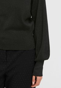 Schwarzer, schimmernder Pullover mit langen Ärmeln, tailliertem Schnitt und voluminösen Ärmeln, kombiniert mit strukturierten schwarzen Hosen mit kleinen gepunkteten Akzenten.