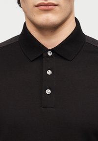 Άντρας που φοράει μαύρο πόλο πουκάμισο με υφή, με τρία ασημένια κουμπιά και διπλωμένο γιακά, εμφανιζόμενος από το πηγούνι έως το στήθος.