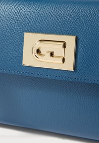 Geantă de mână din piele albastră cu finisaj texturat, având un detaliu logo din metal auriu și cusături vizibile pe margini.