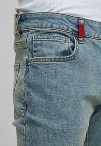 Bližnji posnetek svetlo modrih denim hlač na strani z vidno sprednjo žepom, zankico za pas, oranžnim šivom in rdečo nalepko na pasu.