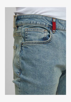 Close-up of light blue denim jeans side with visible front pocket, belt loop, orange stitching, and red tag on the waistband.