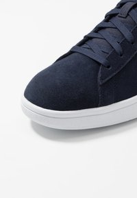 Navy suede sneaker with white rubber sole, featuring flat laces and a rounded toe design; smooth texture with minimal stitching details.